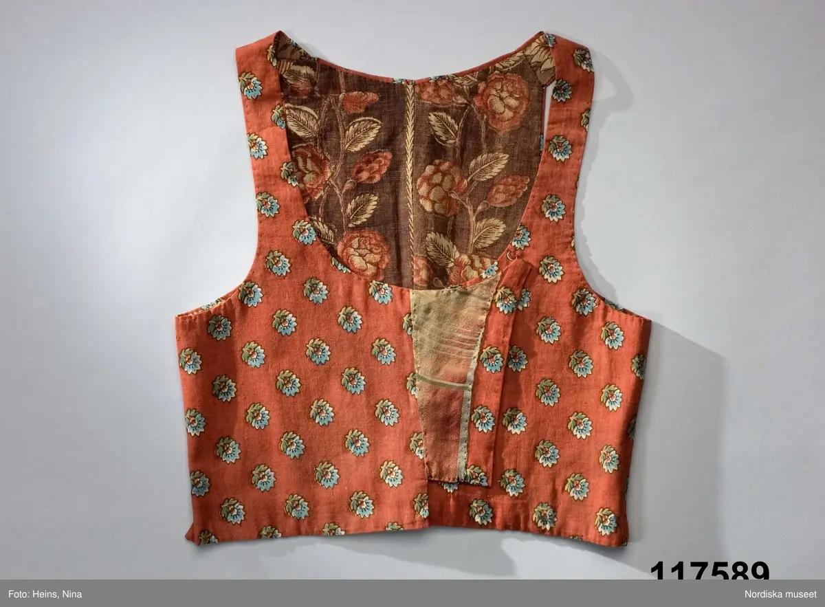 Livstycke av tryckt bomullstyg, kallat kattun, botten färgad i mörk krappröd färg med utsparade små blommor jämnt spridda över ytan. Blommorna har sedan tryckts i med blått och gult med svarta konturer. 2 fram/sidstycken och 4 ryggstycken som avsmalnar ned i ett rudimentärt skört utan veck, små infällda kilar i sidorna. Knäppt med 3 par hakar och hyskor, knäppningen dold under ett förslag av kattuntyget och en bit från ett halskläde i sidensatin med kantbården kvar. Livstycket fodrat med 2 olika kattuntryck av troligen något äldre datum, i ryggen ett brunbottnat bomullstyg med rosenrankor i krapprött och den oblekta grundfärgen, och i framstyckena ett bladmönster i gallerverk i samma färgskala men något ljusare. Troligen är det senare tyget också ett återbrukat halskläde där man skickligt lagt kantbården så att den följer även runt livstyckets kanter.
/Berit Eldvik 2006-12-08