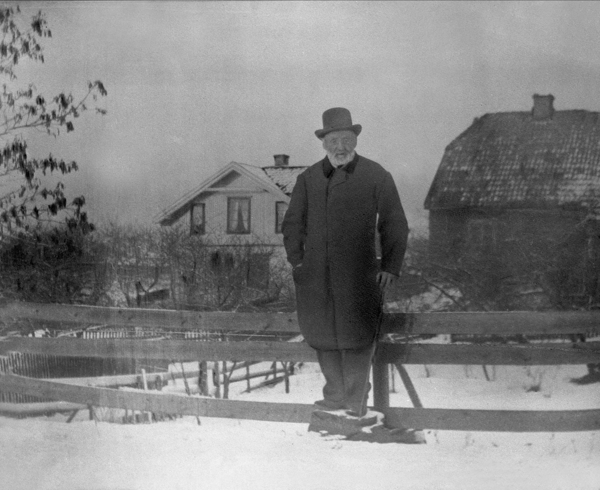 Avfotografering. Eldre mann i yttertøy i hagen, vinter. Våningshus og uthus i bakgrunnen.