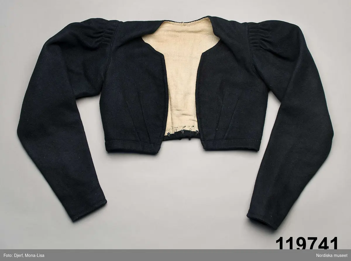 Kvinnotröja av mörkblå vadmal, i modesnitt från 1830-talet. Kort med vida ärmar. 2 framstycken/sidstycken, med 2 sneda insyningar fram upp mot bysten. 3 ryggstycken med svängda sömmar med passepoaler, i nederkant påsydd 2 cm bred kant med litet  rudiment av ryggskört bestående av tre utstående avrundade flikar.  Isydd ensömsärm med mycket vidd upptill samlad i lagda veck på ärmkullen nedsydda med en tvärgående söm 2,5 cm från ärmsömmen, ärmen avsmalnar nedåt  och har vid handleden ett kort sprund knäppt med en hake och hyska. Fodrad med blekt linnelärft, i ärmarna av grövre kvalitet. Knäppt nere på framkanten med 4 hakar och hyskor,  bak i fodret 3 hakar för att fästa mot kjolen.
I mycket gott skick.
Jfr ärmarna med tröja nr 82189 a från Halland. Även detta en folklig anpassning av modedräktens fårbogsärm.
/Berit Eldvik 2010-10-22