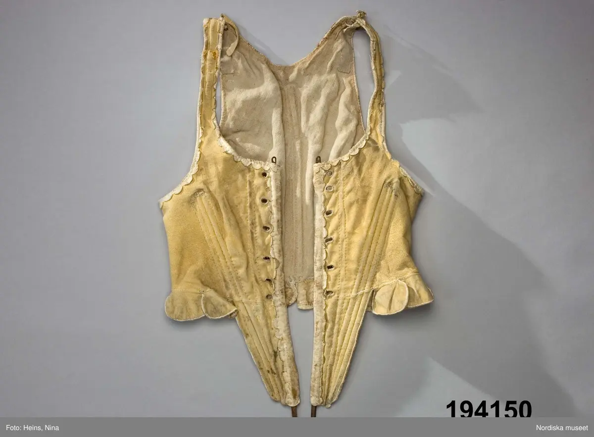 Livstycke av ljust sämskskinn i 1700-talsmodell med djupa snibbar fram styvade med kraftiga järntenar i framkanterna och tunnare träspröt genom sydda diagonala kanaler från brösthöjd och ned i snibbarna, och på motsvarande tre ställen i ryggen. I nederkanten en rad njurformade skörtflikar som överlappar varandra och är kantade med vita skinnremsor. Vita uddstansade skinnremsor även kring framkanter, hals-och ärmringning. Snörning i 6 par kantsydda snörhål. Foder av oblekt linnelärft.
Enligt uppgift i huvudliggaren en fästmöpresent tillverkad av Andreas Andersson i Västergården, Djursätra.
Anm. Järnspröten sticker ut både upptill och nedtill, spridda rostfläckar och allmänt slitet.
/Berit Eldvik 2008-01-22