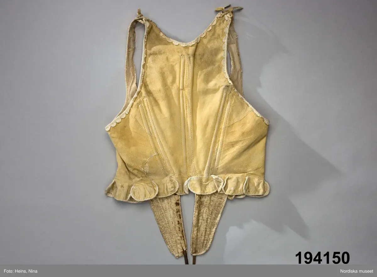 Livstycke av ljust sämskskinn i 1700-talsmodell med djupa snibbar fram styvade med kraftiga järntenar i framkanterna och tunnare träspröt genom sydda diagonala kanaler från brösthöjd och ned i snibbarna, och på motsvarande tre ställen i ryggen. I nederkanten en rad njurformade skörtflikar som överlappar varandra och är kantade med vita skinnremsor. Vita uddstansade skinnremsor även kring framkanter, hals-och ärmringning. Snörning i 6 par kantsydda snörhål. Foder av oblekt linnelärft.
Enligt uppgift i huvudliggaren en fästmöpresent tillverkad av Andreas Andersson i Västergården, Djursätra.
Anm. Järnspröten sticker ut både upptill och nedtill, spridda rostfläckar och allmänt slitet.
/Berit Eldvik 2008-01-22