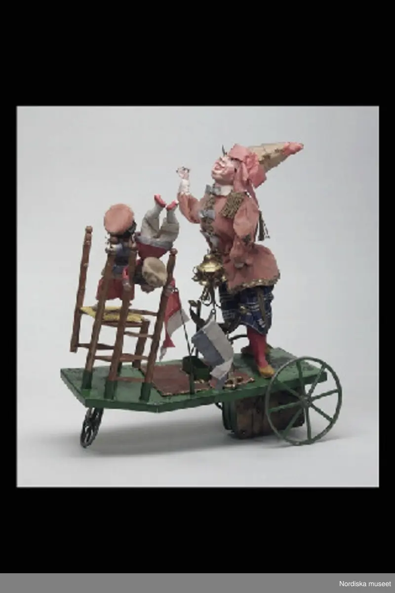 Inventering Sesam 1996-1999:
a+) Mekanisk leksak L 28 cm, B 12,5 cm, H) 45,5 cm
b) Nyckel L 10 cm
c) Originalförpackning L 37 cm, B 17 cm, H 35 cm
a+) Equilibriste ¿ Mecanique (mekanisk balanskonstnär) bestående av clown av massa (papier mach¿?), med rosa sammetsjacka och blårutiga byxor, stående på grönlackerad vagn av metall med tre hjul. På näsan har clownen fäste (spets) på vilken kan fästas. På vagnen olika fästen (bl a två svärdsskidor) för olika lösa attiraljer: Stege med docka i röd sidenjacka och beige huvudbonad, stol (delar saknas) av trä med gul sits och stående på sitsen liten docka med blå jacka, tvådelad flaggstång med grön/vit och röd/vit tvåuddiga flaggor, metallten med klocka och halvmåne i gulmetall med bjällor, Svärd av gulmetall (har ursprungligen trol. funnits två svärd)
Undertill på vagnen finns ett fjäderverk som driver vagnen frammåt och får fästet/spetsen på clownens näsa att snurra. Enligt äldre kat.kort driver fjäderverket även speldosa (?). På spetsen på clownens näsa kan de olika lösa attiraljerna fästas.
b) Nyckel, av vitmetall, ovalt huvud utan hål.
c+) Originalförpackning, enkel, av obehandlat trä, Stor lucka framtill, lock ovanpå. Enligt äldre kat.kort skall det ursprungligen ha suttit ett reklamkort på locket, vilket det nu bara finns reser kvar av.
Har tillhört brukspatron Edv. Otto Benedicks (1818-1872), Gysinge, Gästrikland. 
Leif Wallin feb 1998