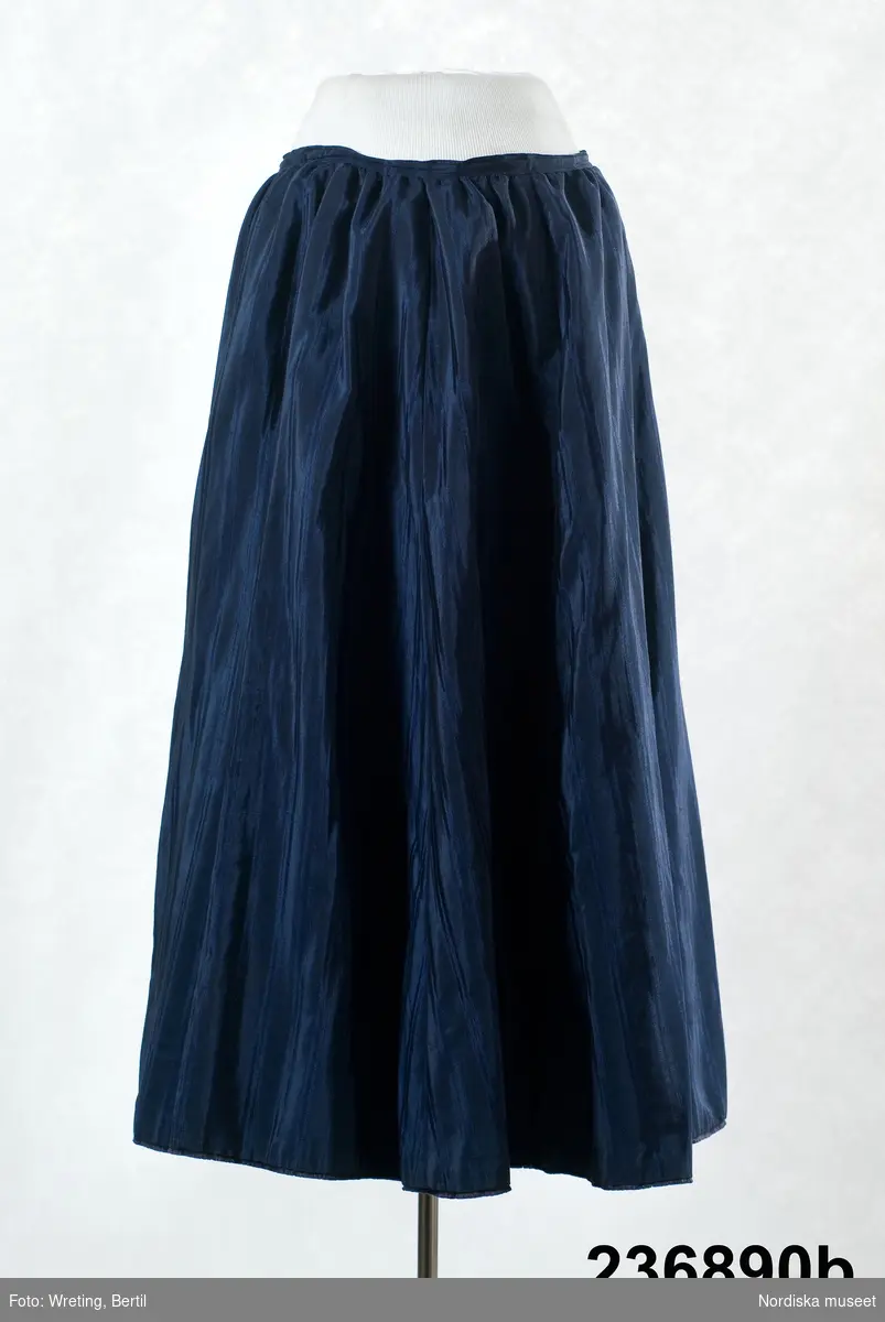 2-delad klänning av mörkblått randvävt halvsiden. a-b samt löst skärp c.

a. Liv figursytt på foderstomme av  ljusgul bomullskypert i fiskbensmönster. 2 framstycken och ett ryggstycke med ett rundat skört bak som dock stoppas ned i kjolen, Fram djupt V-ringad med  2 breda lagda veck som  löper i V-form från axel ned mot underkanten både fram och bak. Längs med vecken även en 3 cm bred  svart maskingjord spets . I ringningen isättning av samma tyg med en mängd tvärgående stråveck, tättslutande ståndkrage  styvad med något mellanlägg. Isydd lång ärm, svängd med 2 sömmar, något rynkad på ärmkullen, snäv vid handleden.
Mitt bak i midjan en fastsydd häkta att fästa mot kjolen. 
b. Kjol av 7 sneddade våder, sydd på foderstomme av svart bomullslärft s.k domestik. Framvåden markerad med nedsydda veck vid sömmarna. Kjolen slät framtill och rynkad bak i midjan mot en 2 cm bred linning av samma tyg. Mitt bak i linningen en fastsydd hyska att häkta mot livet. Dolt sprund i vänster framsöm knäppt med  5 tryckknapapr och överst en hake och hyska. Infälld ficka i sprundet. Skoning i nederkanten av samma tyg som fodret. I kanten slitband med plyschkant.
Anm. 10-12 cm av tyget invikt upptill vid linningen. Kanske ett sätt att inte klippa bort tyg om man skulle vilja återanvända det senare. Det var mycket vanligt att just tygrika kjolar kunde sys om till annat.
c. Löst skärp av samma tyg.  Veckat på längden. Ofodrat, söm mitt bak, 3 fastsydda stålfjädrar på insidan. Knäpps med 3 svarta hakar och tränsade trådöglor 
Klänningen bör vara sydd efter 1903 då Prym uppfann tryckknappen.
/Berit Eldvik 2011-09-08