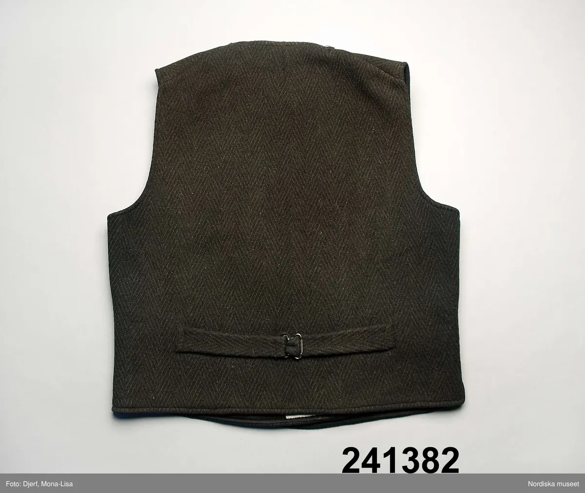 Huvudliggaren (år 1949-1950):
"241382 Storväst, till mansdräkt, av svart ylle i grov spetskypert. Dubbelknäppt med fem knappar. Tre ytterfickor. Foder av randigt, kyprat ylle. Gåva 1950 21/11 Fr. Gustava Nordström Vännäsby. Västerbotten Vännäs sn."

Väst utan krage, dubbelknäppt med gråsvarta fyrhåliga rockknappar, knapphål på båda framstyckena. Tyget mycket kraftigt. 2 framstycken och ett ryggstycke med spänntamp i midjan med svartlackerad metallsölja. Infodringar längs framkanter av tunnare mörkgråmelerat ylle, fodret i yllekypert, bredrandigt i diskret mörkblått och grått på svart botten.  3 iskurna stolpfickor. Västen maskinsydd.
Anm. Något sliten och lagad i fodret uppe i halsen f.ö i mycket gott skick. 
Denna typ av grövre arbets-och vardagsplagg för män var de första som fanns att köpa färdigsydda på landsbygden under 1800-talets andra hälft. De köptes framförallt av män med ambulerande arbeten som skogshuggare, rallare, sjöfolk m.fl. Inga uppgifter finns om just denna väst hör till den gruppen.Se även 241.381. 
/Berit Eldvik 2011-05-13
