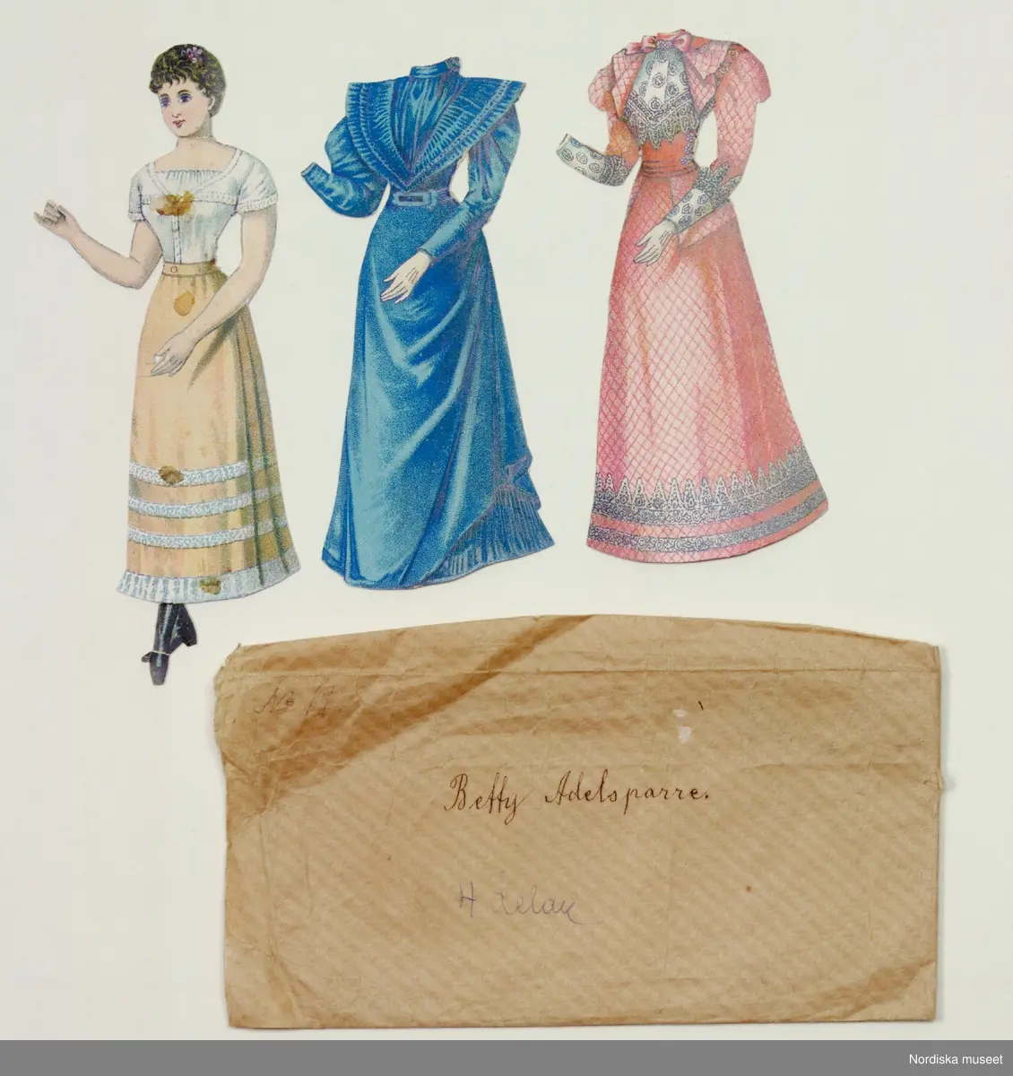 Katalogkort:
"Pappersdocka, dam 1890-tal. Docka i underkläder med två klänningar. I papper märkt Betty Adelsparre.Har tillhört giv:s moster och mor, Sonja och Aina Wistrand, döttrar till chefen för kustbevakningen i Övre Norrland och Göteborg, Helmer Wistrand. BH febr. -68"