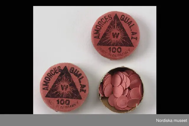 Inventering Sesam 1996-1999:
Diam 3 cm
H 1 cm
2 st små runda askar med lösa lock med nedvikt kant, av kartong, botten grön, locket rött med svart tryck; fylld svart triangel med "W" i mitten i strålkrans. Text "AMORCES QUAL.AI / 100 / MADE IN GERMANY" Båda askarna nästan fulla med röda pappersrundlar med inkapslat explosionsämne.
Hör till knallpulverpistol inv.nr 303.683
Testamentarisk gåva till Nordiska museet och Skansen av hela kvarlåtenskapen, varur valts diverse föremål, möbler, husgeråd, textilier, arkivalier, böcker mm för inmärkning.
Bilagor
1979 TC [=Teje Colling]