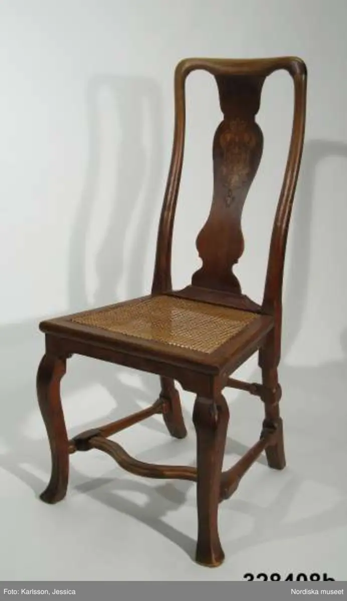 Stol, 6 st (a-f), England, 'Queen Anne', omkring 1720, av valnöt. Hög, balusterformad ryggbricka med inlagd (inkrusterad) dekor av ljusare träslag. Cabriolformade ben och svängt fotkryss med svarvade sidoslåar och bakre tvärslå. Sits med låg sarg, rottingflätad (rottingflätningen nygjord omkring 2002). Tillverkarinitialer inslagna på ben, ryggståndare och baksarg:
a) 'SH' (alt 'HS' ) samt 'IB'.
b) Rund stämpel: [kryss med dubbla streck] 
c) 'SH' (alt 'HS) samt 'IB'
d) 'SH' (alt HS') samt 'IB'
e) 'RH' (3 gånger) 
f) 'RH' (3 gånger),  'IB' (2 gånger) samt 'SH' (alt 'HS')

Jämför stol, inv nr 104.416 med initialerna 'RH' (enligt katalogkort). De engelska stolarnas märkning med tillverkarinitialer på t ex ben, sarg och ryggståndare visar på en rationalisering i tillverkningsprocessen. Initialerna är ibland placerade uppochnedvända vilket indikerar att de slogs in innan stolen sattes ihop. (se 'English furniture 1660-1714 - From Charles II to Queen Anne', Adam Bowett, Italien 2002, s. 264).
Anm: Flera stolar kompletterarade med delar av nytt trä, ilusade bitar. e) bakre tvärslå sekundärt förstärkt med skruv.
/Anna Womack 2008-09-25

Nordiska museet erbjöds 1992 att som testamentarisk gåva efter då framlidna Louise de Kiriline, Ontario, Kanada, ta emot en grupp sittmöbler bestående av två karmstolar och sex stolar. I samtal mellan arvinge till testator och dåvarande avdelningschefen Ingrid Bergman framgick att bakgrunden till gåvan var att del/delar av möblemanget redan skulle finnas i museets samlingar. En från Kanada översänd polaroidbild visade att en eller möjligtvis två stolar av denna typ, troligen tilllverkade i England omkr. 1720, förvärvats av museet redan 1905. Eftersom möbelgruppen skulle ge ett välkommet tillskott av engelska möbler till Charles Totties malmgård på Skansen tackade Nordiska museet ja till den erbjudna gåvan. Stolarna, som vid ankomsten till Sverige visade sig ha sekundära sitsar, har genom konservatorn Sören Lilja vid Skara museum åter försetts med rottingflätade sitsar av ursprunglig typ. Enligt testators anteckningar hade möblerna en gång tillhört den berömda romanförfattarinnan Sophie von Knorring (1797-1848). På sitt fäderne tillhörde hon ätten Zelow, som under 1700-talet haft stark förankring i Västergötland och även på hennes mans sida var det västgötska inslaget påfallande. De båda stolarna som redan tidigare tillhörde Nordiska museet, inv nr 104.415-104.416, inköptes 1905 från Göteborgs Historiska Museum dit de kommit från den kände samlaren Magister Nilsson i Skara. Dessa stolar är av samma typ, men olikheter i detaljer visar dock att de sannolikt ej ingått i samma möblemang. "Magister Nilsson", adjunkt vid Skara läroverk, som var en eldsjäl i Hazelius anda samlade kulturföremål från sin västgötska hembygd och de stora överensstämmelserna mellan dessa stolar och de stolar som nu förvärvas, visar att typen förekommit i Sverige under 1700-talet och kanske särskilt i de västra landskapen kring västerhavsporten Göteborg. Förvärvet behöver därför inte ses enbart relaterat till Tottieska malmgårdens möblering./Per Falck och Birgitta Martinius 2002.

Testator Louise de Kiriline Lawrence (född Flach, 1894-1992), var född i Sverige och växte upp på godset Svensksund på Vikbolandet i Östergötland. Efter att ha arbetat som röda korssyster under 1:a världskriget och i Ryssland efter revolutionen emigrerade hon till Kanada 1927. 
