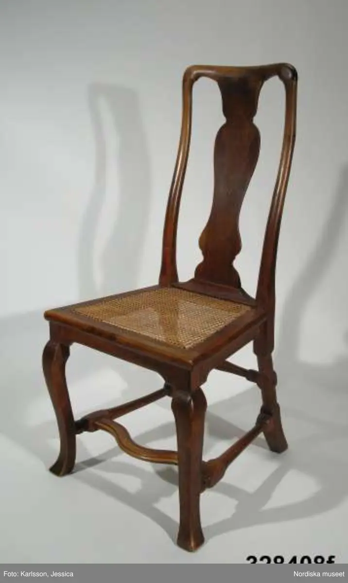 Stol, 6 st (a-f), England, 'Queen Anne', omkring 1720, av valnöt. Hög, balusterformad ryggbricka med inlagd (inkrusterad) dekor av ljusare träslag. Cabriolformade ben och svängt fotkryss med svarvade sidoslåar och bakre tvärslå. Sits med låg sarg, rottingflätad (rottingflätningen nygjord omkring 2002). Tillverkarinitialer inslagna på ben, ryggståndare och baksarg:
a) 'SH' (alt 'HS' ) samt 'IB'.
b) Rund stämpel: [kryss med dubbla streck] 
c) 'SH' (alt 'HS) samt 'IB'
d) 'SH' (alt HS') samt 'IB'
e) 'RH' (3 gånger) 
f) 'RH' (3 gånger),  'IB' (2 gånger) samt 'SH' (alt 'HS')

Jämför stol, inv nr 104.416 med initialerna 'RH' (enligt katalogkort). De engelska stolarnas märkning med tillverkarinitialer på t ex ben, sarg och ryggståndare visar på en rationalisering i tillverkningsprocessen. Initialerna är ibland placerade uppochnedvända vilket indikerar att de slogs in innan stolen sattes ihop. (se 'English furniture 1660-1714 - From Charles II to Queen Anne', Adam Bowett, Italien 2002, s. 264).
Anm: Flera stolar kompletterarade med delar av nytt trä, ilusade bitar. e) bakre tvärslå sekundärt förstärkt med skruv.
/Anna Womack 2008-09-25

Nordiska museet erbjöds 1992 att som testamentarisk gåva efter då framlidna Louise de Kiriline, Ontario, Kanada, ta emot en grupp sittmöbler bestående av två karmstolar och sex stolar. I samtal mellan arvinge till testator och dåvarande avdelningschefen Ingrid Bergman framgick att bakgrunden till gåvan var att del/delar av möblemanget redan skulle finnas i museets samlingar. En från Kanada översänd polaroidbild visade att en eller möjligtvis två stolar av denna typ, troligen tilllverkade i England omkr. 1720, förvärvats av museet redan 1905. Eftersom möbelgruppen skulle ge ett välkommet tillskott av engelska möbler till Charles Totties malmgård på Skansen tackade Nordiska museet ja till den erbjudna gåvan. Stolarna, som vid ankomsten till Sverige visade sig ha sekundära sitsar, har genom konservatorn Sören Lilja vid Skara museum åter försetts med rottingflätade sitsar av ursprunglig typ. Enligt testators anteckningar hade möblerna en gång tillhört den berömda romanförfattarinnan Sophie von Knorring (1797-1848). På sitt fäderne tillhörde hon ätten Zelow, som under 1700-talet haft stark förankring i Västergötland och även på hennes mans sida var det västgötska inslaget påfallande. De båda stolarna som redan tidigare tillhörde Nordiska museet, inv nr 104.415-104.416, inköptes 1905 från Göteborgs Historiska Museum dit de kommit från den kände samlaren Magister Nilsson i Skara. Dessa stolar är av samma typ, men olikheter i detaljer visar dock att de sannolikt ej ingått i samma möblemang. "Magister Nilsson", adjunkt vid Skara läroverk, som var en eldsjäl i Hazelius anda samlade kulturföremål från sin västgötska hembygd och de stora överensstämmelserna mellan dessa stolar och de stolar som nu förvärvas, visar att typen förekommit i Sverige under 1700-talet och kanske särskilt i de västra landskapen kring västerhavsporten Göteborg. Förvärvet behöver därför inte ses enbart relaterat till Tottieska malmgårdens möblering./Per Falck och Birgitta Martinius 2002.

Testator Louise de Kiriline Lawrence (född Flach, 1894-1992), var född i Sverige och växte upp på godset Svensksund på Vikbolandet i Östergötland. Efter att ha arbetat som röda korssyster under 1:a världskriget och i Ryssland efter revolutionen emigrerade hon till Kanada 1927. 
