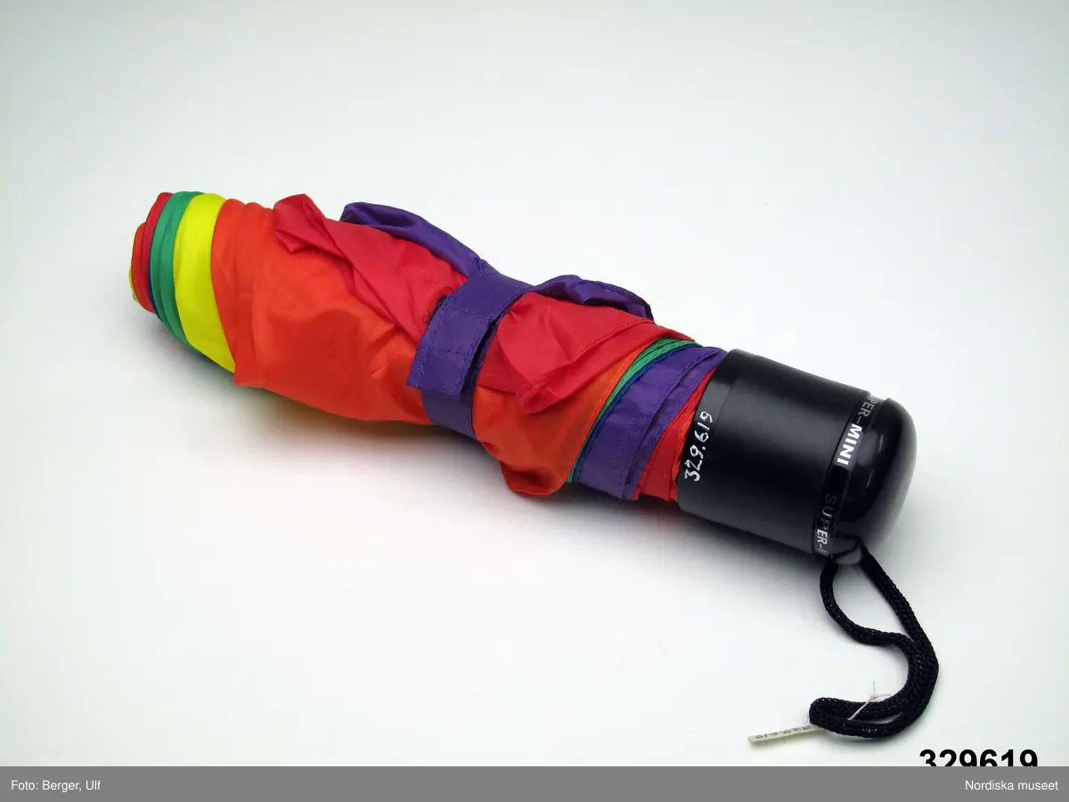 Paraply med svart plasthandtag med svart snodd i konstmaterial. Ställningen i vitmetall. Paraplytyget av nylon i olika tårtbitsformade fält i färgerna blått, lila, två nyanser rött, gult och grönt. På handtaget påklistrad etikett med texten "SUPER-MINI". Litet band av nylon fäst på det lila fältet. Bandet, som är försett med kardborreband, sätts runt paraplyet i ihopfällt tillstånd för att hålla tyget på plats. 

Paraplyet köptes av givaren och hans pojkvän under Stockholm Pride / EuroPride 2008 vilket är den största årliga festivalen för HBT-personer i Sverige och 2008 firades 25 juli-3 augusti.

Utdrag ur intervju med givaren: De köpte tre likadana paraplyer i QX shop i Pride Park. Pojkvännen tyckte att de kunde vara bra att ha i den stora paraden på lördagen som de deltog i. Det regnade en del under de sista dagarna under Prideveckan och speciellt på lördagen innan och under paraden. Ett regnbågsfärgat paraply tyckte de var bra då det tydligare visar att man är en HBT-person. Veckobandet och dogtagen syns ju inte på lite längre avstånd. Givaren tror att paraplyerna kostade 99kr/st vilket han tycker är ganska mycket för ett så litet och enkelt paraply och då liknande paraplyer brukar kosta mindre. Men eftersom det var regnbågsfärgat så tyckte han det var värt det i alla fall. 
De hade tänkt använda paraplyerna som vanliga paraplyer efter Prideveckan, men ett har de lånat ut och inte fått tillbaka, och ett har redan gått sönder och det tredje är detta som museet får. 

Givaren och hans pojkvän, som har varit på de flesta Pride-firanden sedan 1998 och de brukar försöka gå på så mycket av aktiviteterna som möjligt under veckan. Se vidare intervju med givaren om hans pridefirande i arkivet (D.457).

Föremålet ingår i Nordiska museets dokumentation av Europride 2008 med föremål, intervjuer och fotografier. Se arkivet D.457. Övriga föremål med anknytning till Peter Csaszti se inv.nr 329.615-329.620 samt 329.669-329.670.
/Leif Wallin 2009-03-06