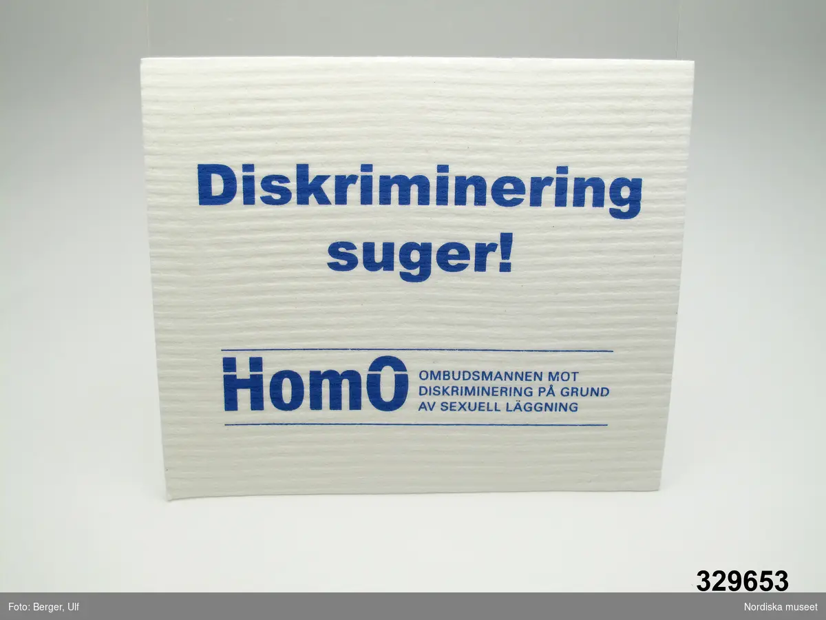 Disktrasa av textil. Rektangulär, vit med blå tryckt text "Diskriminering suger!" och "HomO OMBUDSMANNEN MOT DISKRIMINERING PÅ GRUND AV SEXUELL LÄGGNING".
/Leif Wallin 2009-06-22