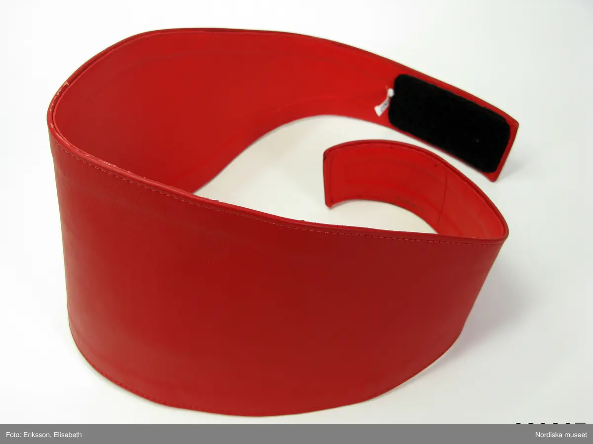 Cummerbund (maggördel) i rött skinn. Två ovala skinnbitar lagda mot varann och fastsydda med maskin. Avigsidans skinn klarare i färgen. Fästs med hjälp av svart kardborrband.

Gördeln uppsydd för Nordiska museet i mars 2009, beställare Peter Siepen. Siepen har själv en snarlik maggördel, men eftersom han inte ville skänka denna till Nordiska museet beställde han en ny från Läderverkstan, Rosenlundsgatan 30A i Stockholm, som museet sedan kunde förvärva. Gördeln användes bl.a. som en del av den outfit som bars av Siepen vid evenemanget Tomterock, vilket är en årligen återkommande "kändisfest" på hotell Amaranten i Stockholm, i november 2008. Hela denna outfit (se inv.nr 329.799-329.808) förvärvades av Nordiska museet i mars 2009, som en del i projektet "Dagens Dandy". Se vidare arkivet D 460.
/Zingoalla Rosenqvist 2009-03-19