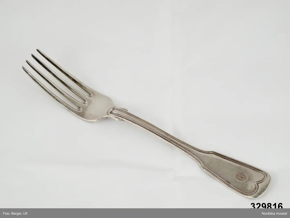 Fyrkloig matgaffel, nysilver, dubbelräffla, hjärtlikt svängd vid rund ända. Modellen är det som senare kom att kallas "Gammalsvensk".

Monogram på framsidan. Klack vid roten. Stämpel på baksidan: N2 -  Zethelius. Förnamnsbokstaven J sammanfallande med första H-stapeln, J-prick ovanför stapeln.

Har tillhört Lärftskramhandlare Johannes Hazelius, Stockholm. Se även inv.nr. 329.815 (stämpel)  och 329.817 (gaffel märkta med denna stämpel).
/2007-03-19 Heidi Henriksson