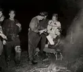 Nattorientering, män och en kvinna i mörker står runt en man som sitter på en fällstol intill en eld. Nordiska Kompaniet 1940.