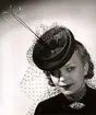 Hattmode visas på Nordiska Kompaniet 1941. kvinnlig modell i liten rund hatt med flor och upprättstående dekoration. I halsen bär hon en brosch med stenar.