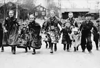 Barn, små kullor och masar, i folkdräkt springer på väg mot fotograf. Barnen kallar till Valborgsmässa 30 april 1936 i Floda genom att ringa i koskällor.