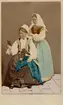 Dräktdockor med huvuden skulpterade av C A Söderman, visade på världsutställningen i Paris 1867. Två kvinnor i folkdräkter från Blekinge, varav en hjälper den andra med huvudbonaden. Handkolorerad version.