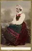 En kvinna i folkdräkt sitter vänd i profil. Högtidsdräkt från Österåker, Södermanland. Som den bars omkr. sekelskiftet 1900
