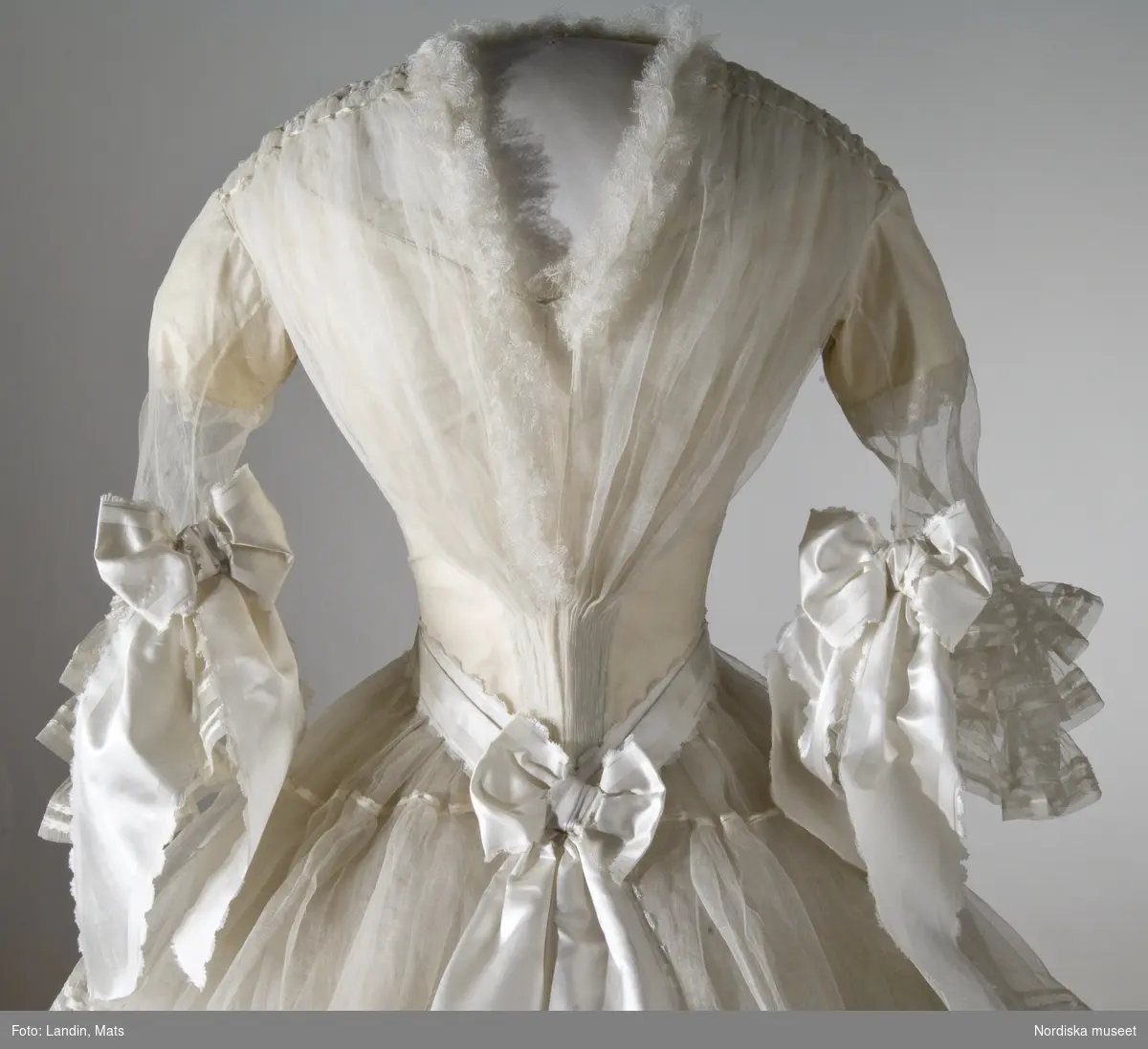 Brudklänning, liv med fastsydd kjol, vit tarlatan, garnerad med vita sidenband, rosetter. Klänningen har ett löst skärp av vit tarlatan.