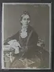 Porträtt av Eugénie, Prinsessa av Sverige och Norge (1830-1889), Sittande knästycke, kroppen vriden åt höger, ansiktet närmast en face. Mittbena, slätt hår kammat bakom öronen, samlat till flätad uppsättning ovanpå hjässan. Mörk krinolin med vertikala snörmakeriband framtill och kring de vida ärmarna. Spetsmanschetter och spetskrage, på kragen oval brosch med kors. Prinsessans h. arm vilar på litet bord, v. arm böjd framför kroppen, händerna hålls intill varandra vid bordsskivans kant.