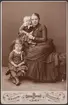 Kabinettsporträtt av kvinna (Henrietta Maria Carlson f. Tempelman f. 7/6 1860 d. 23/6 1950) gift med Fredrik Carlson (1854-1927) med två barn (kapten Arne Carlson f. 20/10 1881 d. 17/9 1921 och musikdirektör Sigrid Carlson f. 9/12 1883 d. 9/7 1935). Kvinnan möjligen sorgklädd bärande sorgsmycken i form av grovt halsband och armband.
Grupporträtt.