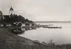 Rättviks kyrka i Dalarna. Kyrkbåtar på Siljans vatten. Vid stranden framför kyrkstallarna står högtidsklädda människor.