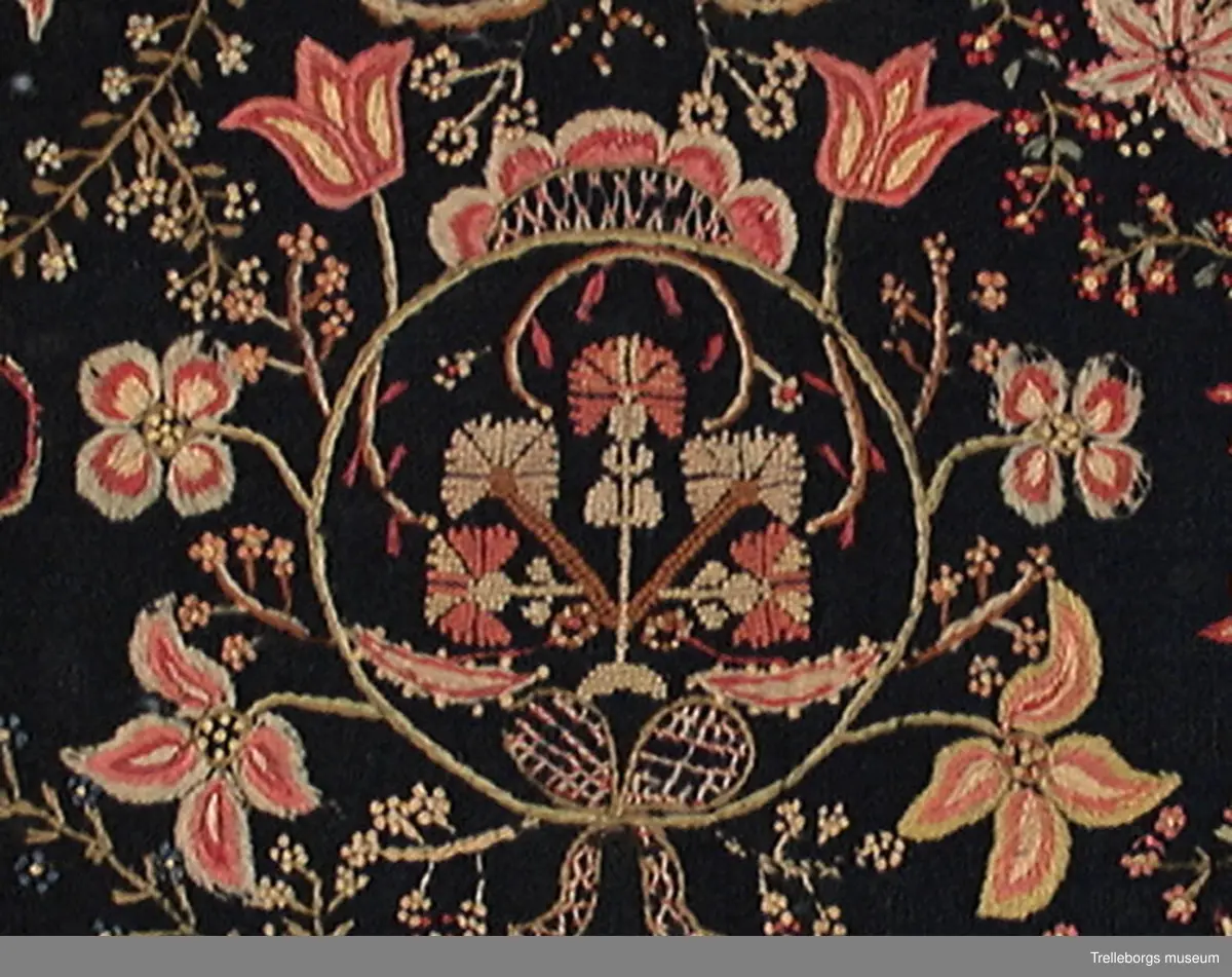 Botten svart ylletyg i tuskaft, broderi: 2 tr ullgarn. Del av åkdyna i plattsömsbroderi, schattersöm. Dynans sirliga rikt utformade mönster är mycket ovanligt. Färgerna är ljusa i rött, rosa, blått och gult. På mitten är en rund ring broderad, denna har en mittfigur sydd med små korsstygn. Runt ringen finns blommor typiska för schattersöm. I varje hörn finns tulpanliknande blommor omgivna av en halv krans småblommor. Småblommorna förekommer över hela ytan. Blomsterbuketten i mitten är sydd i små korsstygn, rosetten i bottensöm, de mindre blommorna är sydda med knutar. Kantad med tyg. Endast halva dynan. Se även Årsbok för Malmö museum sid 44-45, Ernst Fischer: I Trelleborgs museum finnes hälften av en ursprunglig åkdyna som har ett mittmönster av en ring-krans med ändarna nedtill sammanknutna av ett band med ögla samt krönt av ett ornament. Från kransen utgår upptill två tulpaner, på mitten två fyrbladiga blommor, och nederst två utslagna tulpaner. Mellan dessa blommor små kvistar. Inom kransen en växt med fem nejlikor sydda i korssöm. I övre hörnet en växt med fem nejlikor sydda i korssöm. I övre hörnet en växt med sexbladiga blommor och i det nedre en tulpanstjälk med tre blommor. Båda omramade av en spröd kvist. Till vänster om kransen upptill och nedtill hälften av en urna med tre stora blommor, vända mot varandra. Även dessa omramade av liknande kvistar.
