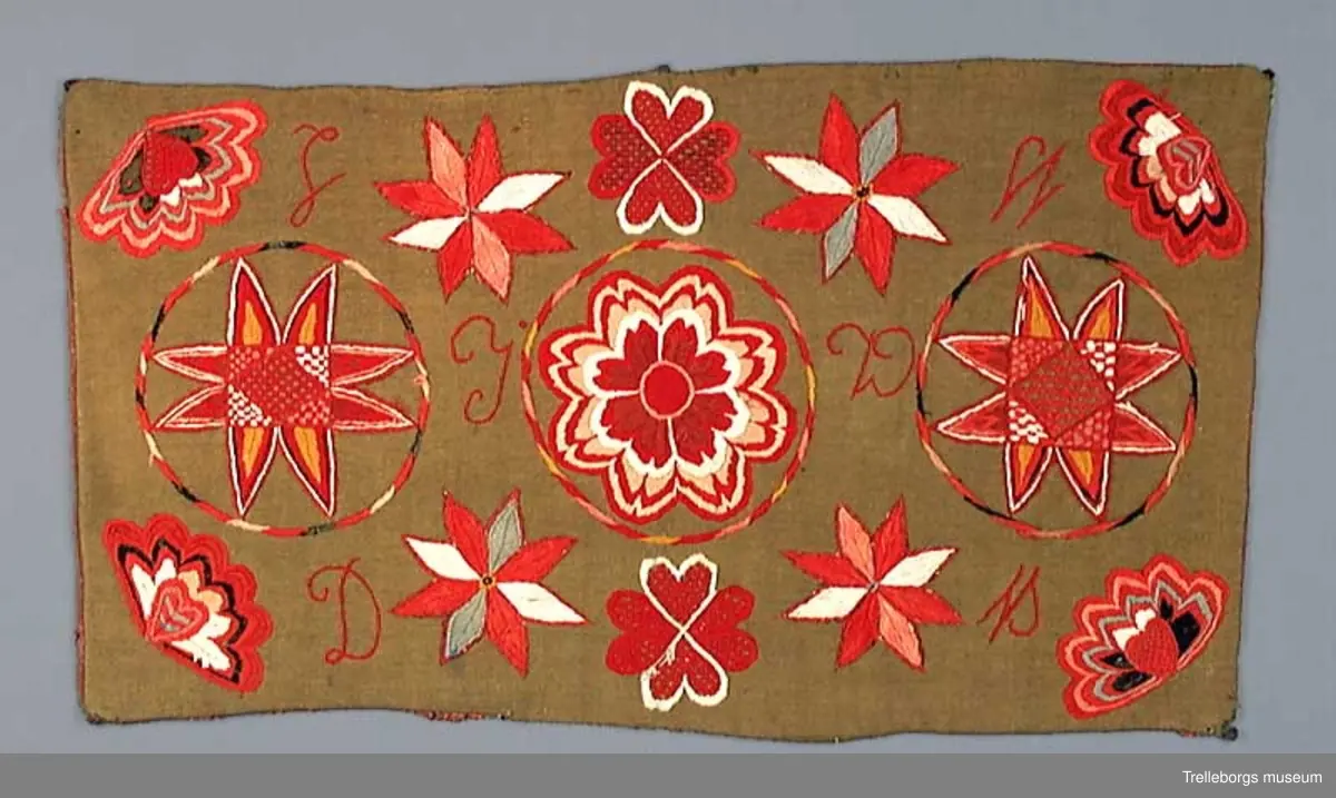 Bottentyg är grön vadmal. Tre stora ringar i kontur utgör huvudmotiv. I ringen i mitten är en stor blomma med 8 kronblad utfyllnad. I de båda ringarna vid sidan är broderat en spetsuddig stjärna med 8 vingpar. Dess centrum är en snedställd kvadrat, sydd i gallerliknande konstsöm. Utmed agedynans långsidor är broderat olika blomsterstjärnor, pionliknande blommor och en blomliknande rundning av 4 st hjärtfigurer. Färger: vitt, gult, svart, blågrått samt många olika nyanser i rött. Baksidan tegelröd yllevävnad. 
Enligt katalogen märkt I.W.D.S.  SL. Hjärtrosetter och hjärtan. 
Enligt Ernst Fischer, Årsbok från Malmö museum 1971, sid 180: En åkdyna i Tbgs museum har som mittmotiv en runduddig stjärna, omgiven av en ring samt en hjärtrosett över och under, flankerad av spetsuddiga stjärnor. T.h. och v om mittmotivet stora spetsuddiga stjärnor inom ringar. I hörnen varierande blomformer. De ganska svårtydda initialerna skulle kunna tydas som LWD IVH, ehuru bokstäverna då icke är placerade i rätt ordning. IVH skall i så fall beteckna I Vämmenhög.