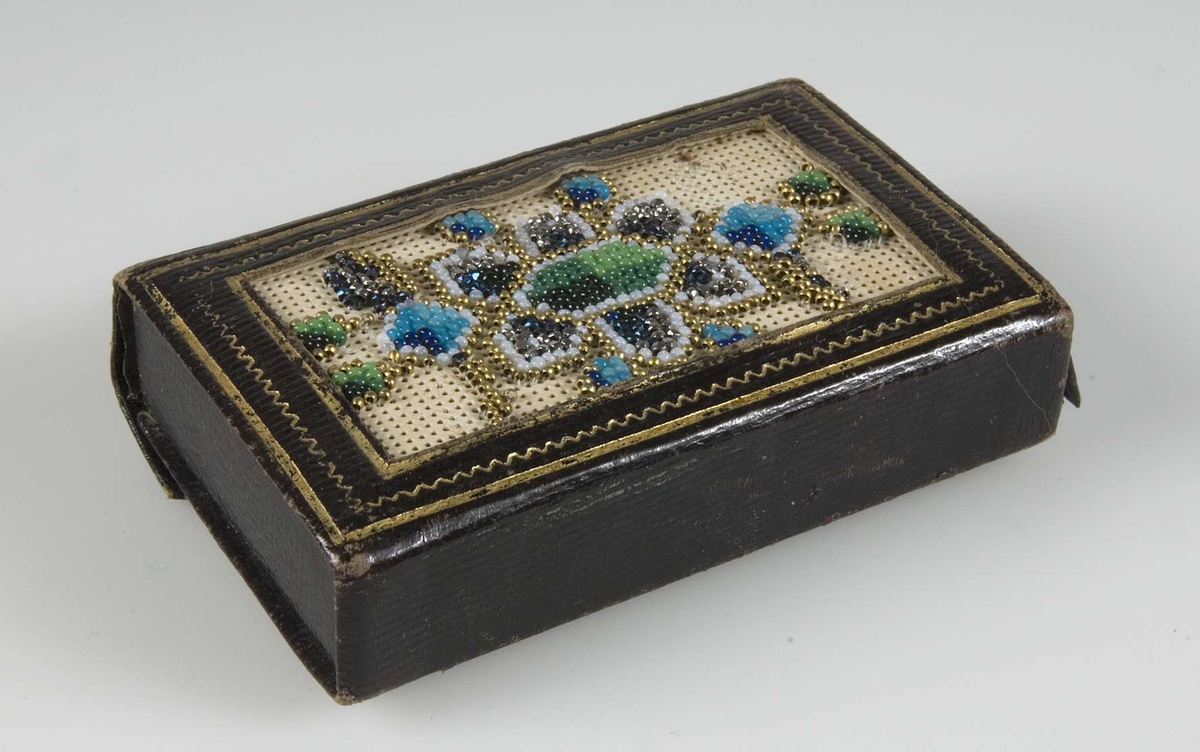 Ask av brunt läder med framsida av pärlbroderi på perforerad papp. Pärlor i grönt, blått, vitt, silver och guld. Askstomme av plåt.
