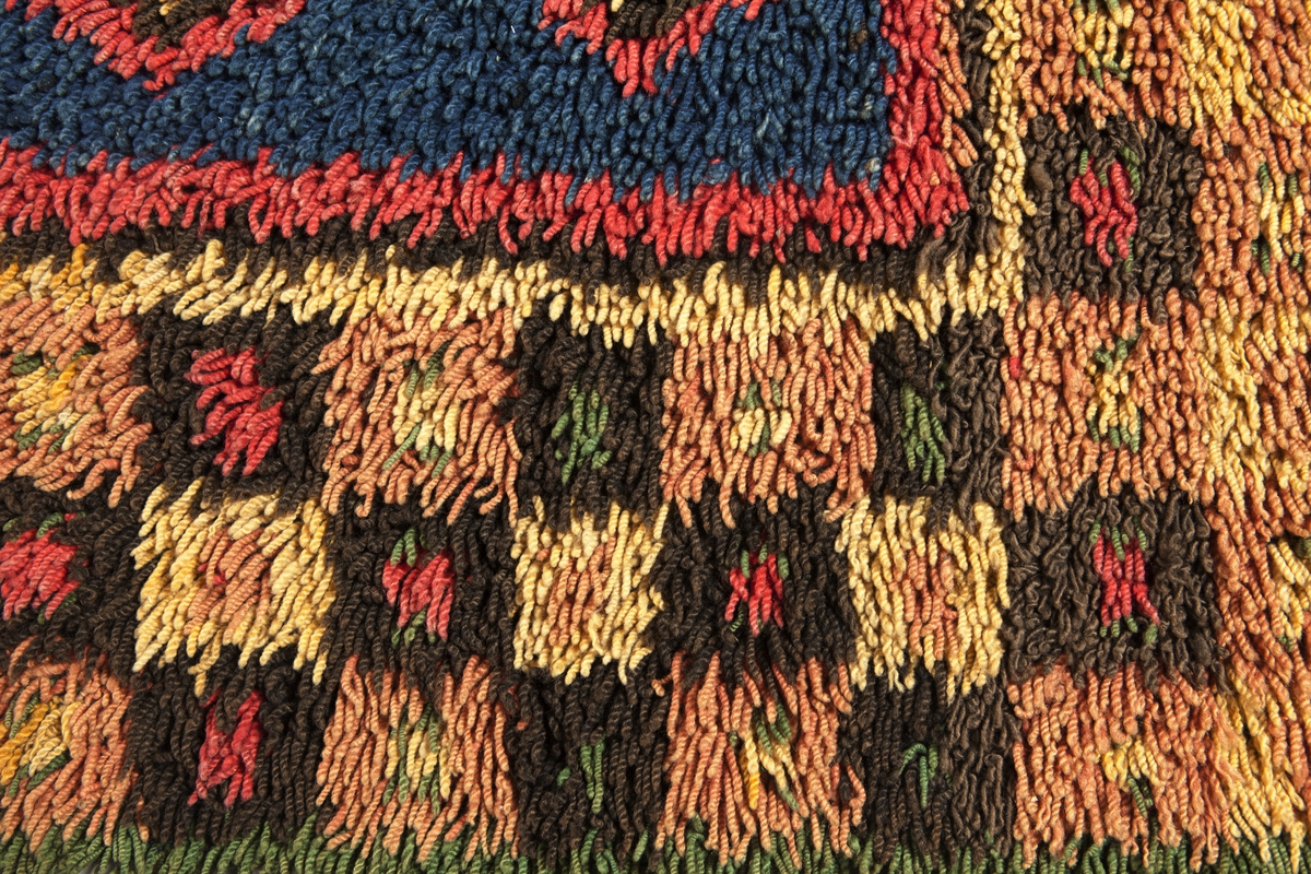 Prydnadsrya i två våder, hopsydda kant i kant. Botten i korndräll med varp av blått s-tvinnat lingarn 11 tr/cm och inslag av mörkbrun s-spunnen ull 7½ inslag/cm. Nockor av en tråd tvåtrådigt z-tvinnat hårdtvinnat ullgarn, knutna med giordesknut över 4 varptrådar med 4 varptrådar mellan nockorna och 4 inslag mellan nockraderna. Nockorna syns ej på baksidan.

Mönster: Stort blått mittfält avgränsat mot bården av en röd, en grönbrun och en gul smal linje. På det blå mittfältet, utmed sidorna romber av rött, grönbrunt och gult. Bred bård av schackrutor i grönbrunt, gult och rosagult. Gröna, röda och gula prickar i rutorna. Ytterst smal grön rand.