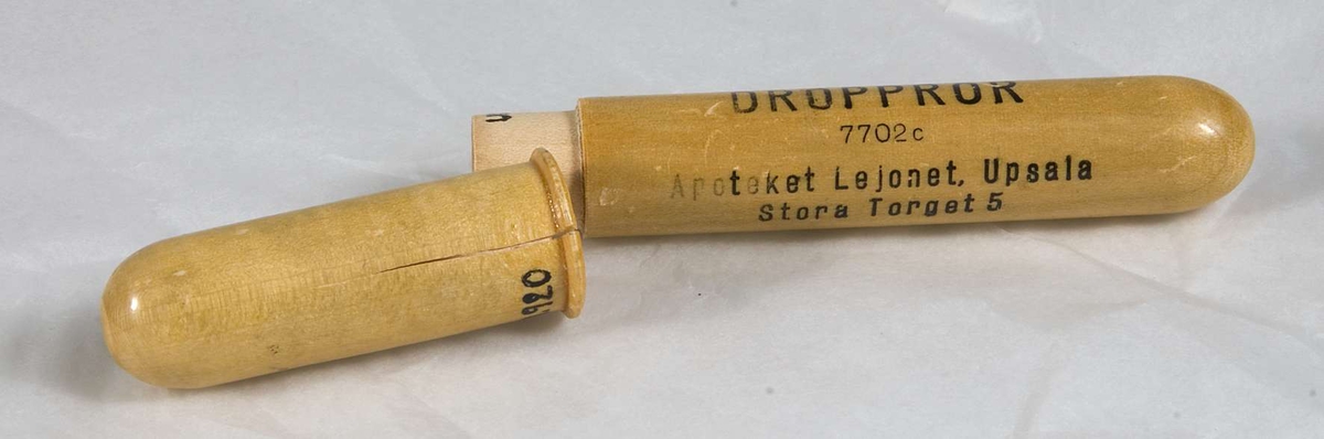Förpackning för dropprör. Rörformigt i ljust lackerat trä. Två stycken som träs i varandra. Ett tryckt lejon i svart. Därunder i svart tryckt text: DROPPRÖR 7702 c, Apoteket Lejonet, Upsala. Stora Torget 5. 