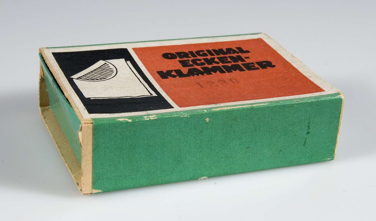 Skjutask av papp klädd med grönt papper. På ovansidan pappersetikett i rött och svart med text: ORIGINAL ECKEN-KLAMMER, 1280 och bild av hur en klämma viks runt hörnet på ett papper.
Innehåller mässingsbleck med vikmarkeringar och räfflor, se UM22320b.
