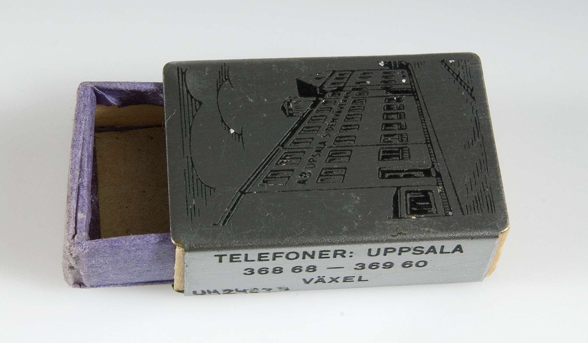 Liten tändsticksask av träspån med hölje av graverat stål, asken klädd med blått papper, bild av AB Upsala Sidenväveriers fabrik på Kålsängsgränd 18, Uppsala, samt reklamtext.