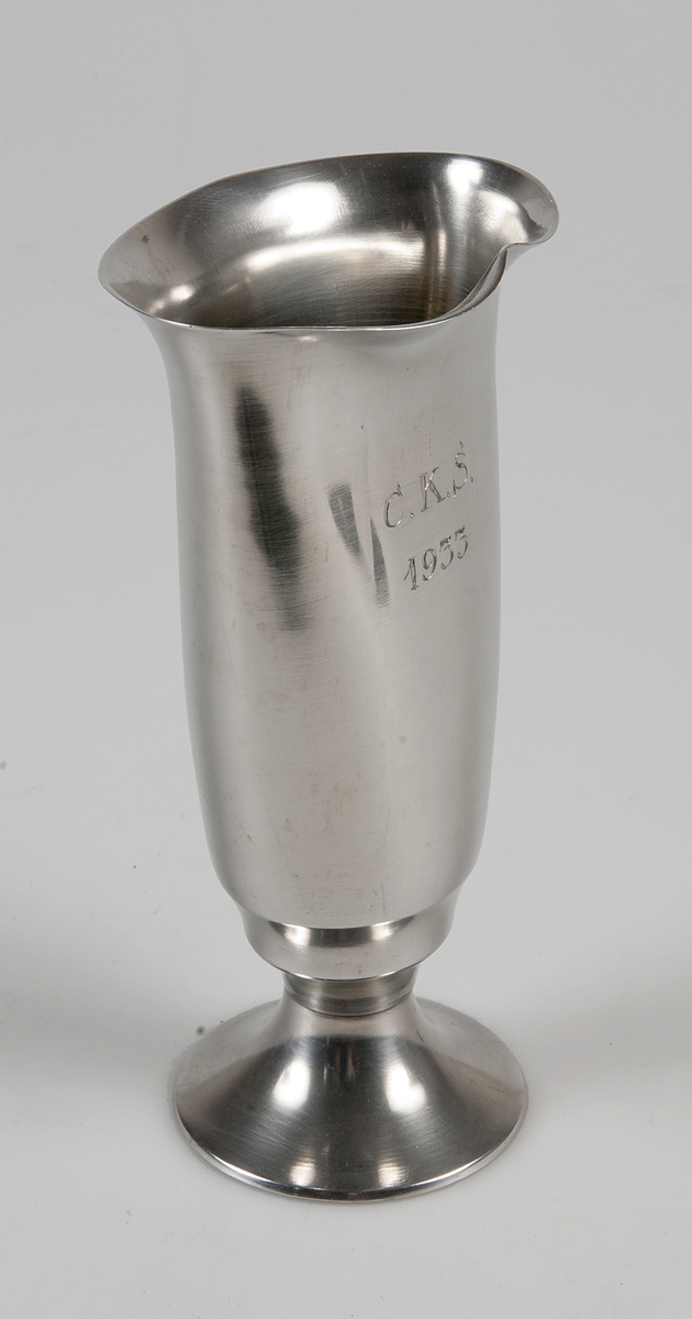 Metallbägare med fot. Smal kuppa, slät med ornament mitt på. Ingraverat: C. K. S. 1933.