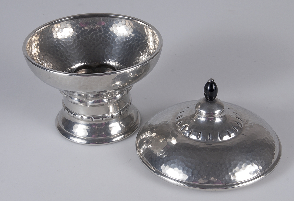 Skål med lock av hamrat tenn. Locket har en svart knopp, eventuellt av trä. Skålen har dekorerat fotparti. Stämplad: MAGS TIN. Ingraverat: Intresserad Thorares Hederspris 1927.

