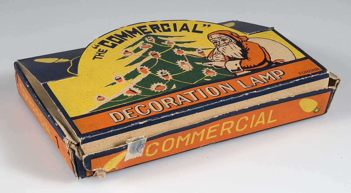 Pappask innehållande kulört elektrisk julgransbelynning, 16 små äggformade lampor av glas i blått, gult, vitt , rött, grönt och orange , i hållare av grön plast och med sladdar klädda med grön tråd. Några lampor är större än de andra och troligen utbytta. På askens lock bild av tomte och julgran i rött, gult, grönt och vitt och text: The Commersial Decoration lamp. Foreign.