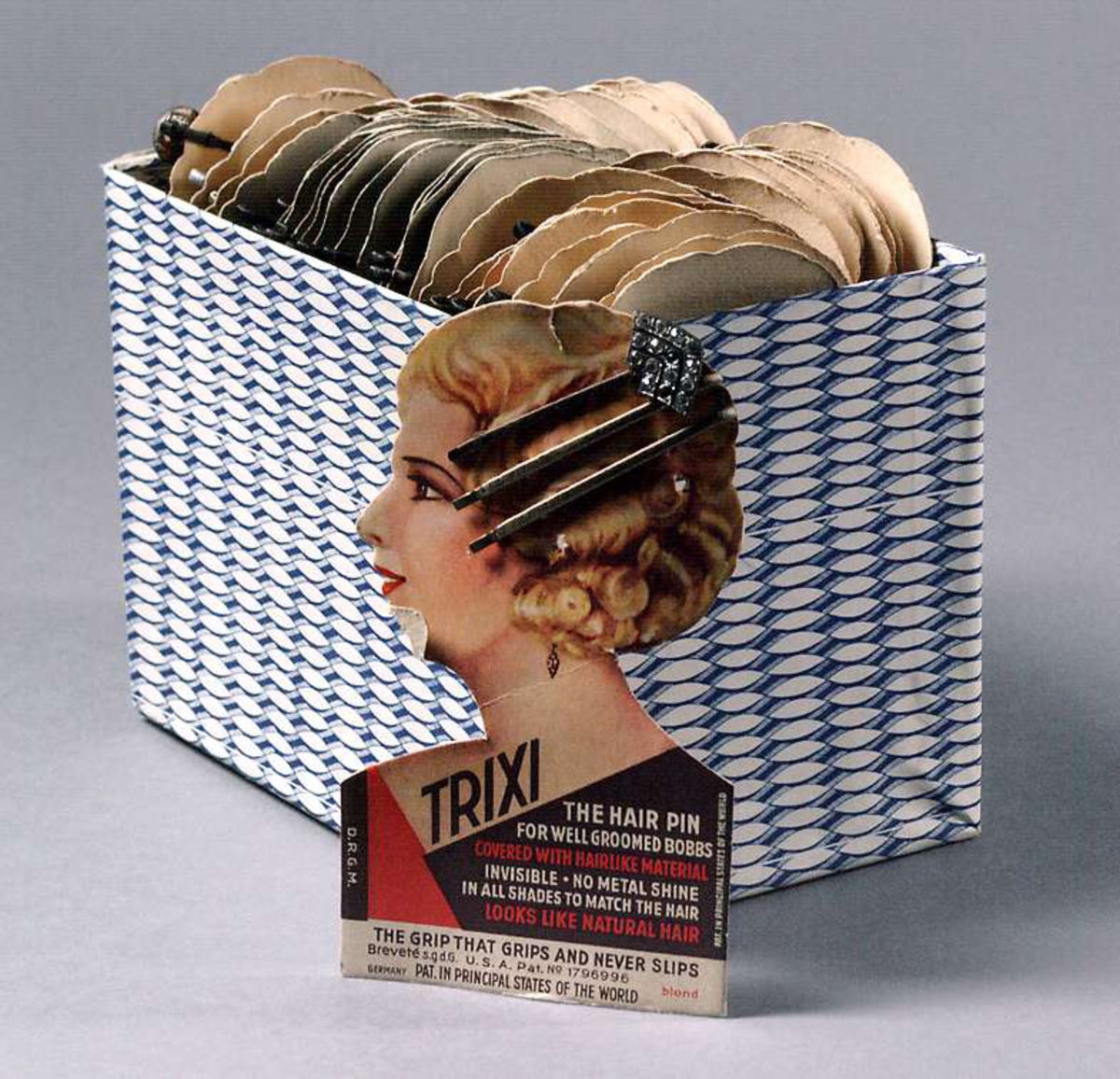 48 förpackningar med två - tre smala klädda hårspännen i varje förpackning. Förpackningarna formade till kvinnoansikte. Tryckt text: TRIXI THE HAIR PIN. Förpackningarna förvaras i blåvit ask. Tillverkade i USA. På baksidan av varje förpackning stämplat 30 resp. 40 öre. Två förpackningar har hårspännen av strass.