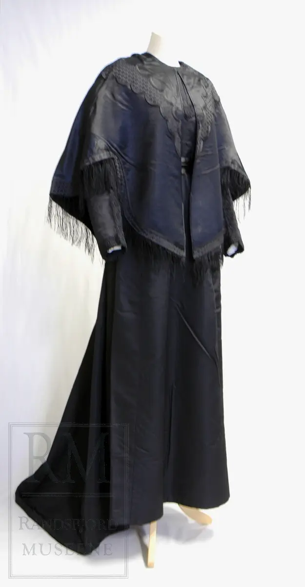 Håndsydd. Svart silkesatin med glanset bakside. Alpakkarenning (?) m / innslag i natursilke. Applikasjon av svart mønstret silkerips. To agramanrosetter med svarte glassperler. Sammensydd i rygg. Lukkes med hekte og malje ved halsåpning foran. Frynser.