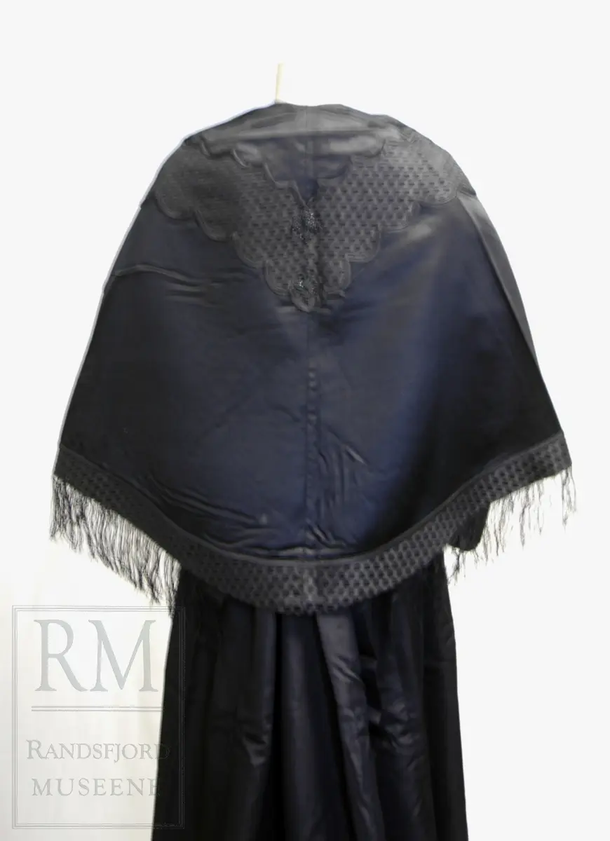 Håndsydd. Svart silkesatin med glanset bakside. Alpakkarenning (?) m / innslag i natursilke. Applikasjon av svart mønstret silkerips. To agramanrosetter med svarte glassperler. Sammensydd i rygg. Lukkes med hekte og malje ved halsåpning foran. Frynser.