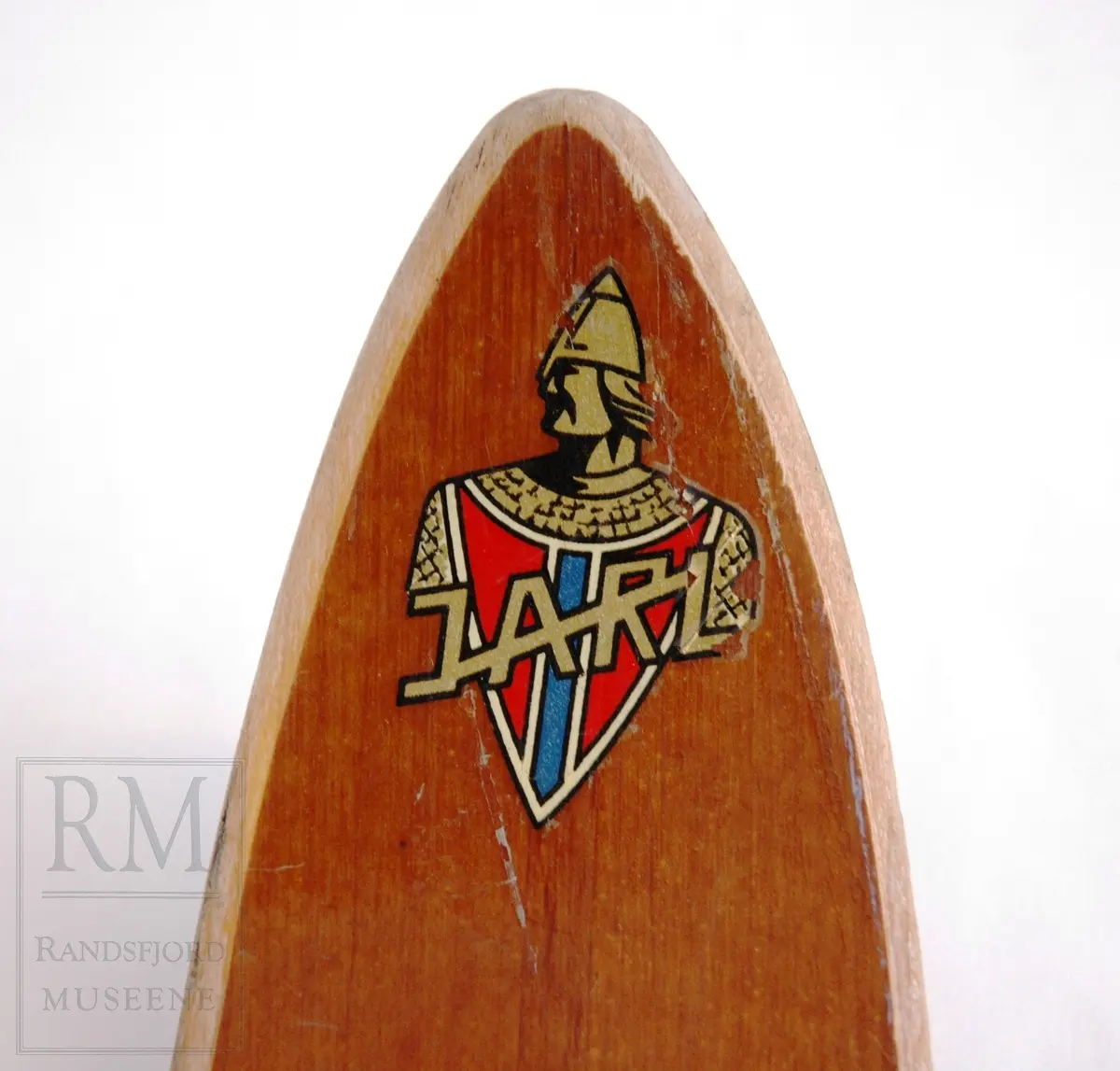 Brunlakkerte treski med fjørbindinger ("Kandahar løypebinding") der det går en fjær bak hælen. Skiene er norskprodusert og av merket Jarl. Varemerket er på begge tupper- en viking i profil med brynje og norske farger.
