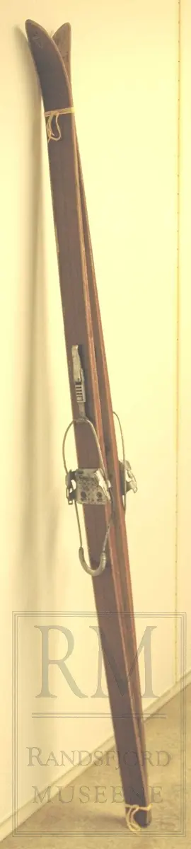 Brunlakkerte treski med fjørbindinger ("Kandahar løypebinding") der det går en fjær bak hælen. Skiene er norskprodusert og av merket Jarl. Varemerket er på begge tupper- en viking i profil med brynje og norske farger.