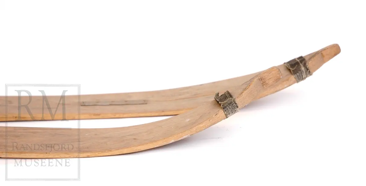 1 par ski, hoppski. Hickory, korkenebb spiss, lyse brune. Tre renner. Tjærebånd på tuppene. Påskrudd metalltåbinding og feste for Kandahar fjærbinding (mangler). Sprukne, reparert med metallplate