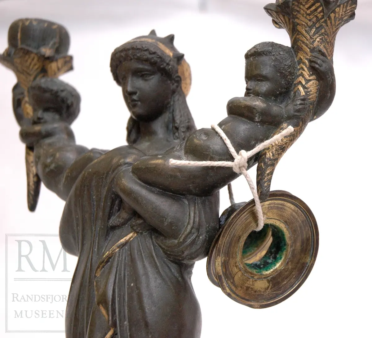 Kvinnefigur på sokkel, med ett barn i hver arm. Barna holder hvert sitt horn (?) med lyseholder. Noe forgylling. Løse lysmansjetter. b) mangler sokkel.