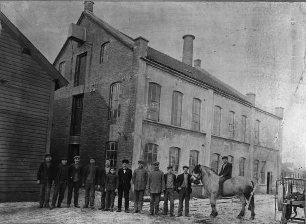 Enok Berge sine arbeidsfolk utenfor fabrikken. F.v. Kristian Sømme, Gabriel Olsen, Bernhard Mikalsen, Johan Rasmussen, Tønnes Rægevig, Harald Tjølsen, Enok Berge, Sigurd Berge, Lukas Berge, Bjarne Ikdahl, Knud Rovig og Karsten Petersen (på Borken).

Supplerande opplysningar frå informant:
Sandnes Dreieri. Innehaver Enok Berge. St. Olavs gt. 20. Bygd ca.: [årstal manglar]
Bildet er med i Dreyer: Sandnes - bilder fra gamle dager, side 138.