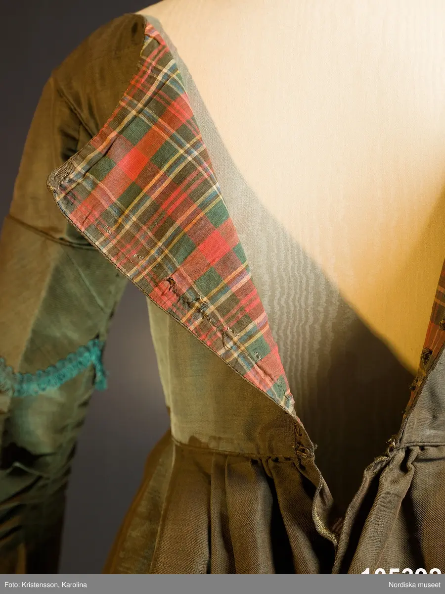 Hel klänning i 1830-talets modesnitt, av fint halvylletyg i kypert med varp av ljusbrunt bomullsgarn och inslag av grönt tunt glansigt redgarn som ger en grönskimrande yta. Livet går ned nästan till midjehöjd, figursytt med 2 framstycken med bakåtflyttade sidsömmar, ett ryggstycke med 6 inprovningar fördelade i två grupper. Vid båtringad halsringning, knäppning med tätt sittande hakar och hyskor. Isydda långa ärmar svängda med 2 sömmar  med 3 veck vid armbågen. Ärmsprund knäppt med hyska och hake samt 4 cm uppslag med rundade hörn. Upptill på ärmen besättning av tyget med stora rundade uddar kantade med klargrön bomullsfrans.  Passepoaler av tyget i alla sömmar. Livet fodrat med skotskrutig bomullslärft i rött, grönt, gult och vitt och i ärmarna ett smalrandigt bomullstyg i blått och rosa. Kjolen sydd av  5 våder, lagda veck runtom i midjan. sprund mitt fram. Uppsytt veck  en decimeter från fållen. Skoning med brun bomullslärft. Handsydd.
Har burits 1838 som ungmorsklänning, alltså av bruden dagen efter bröllopet, av Maria Margareta Mårtensdotter från Kysings, Vall sn på Gotland. Möjligen tillsammans med sidenhalsklädet 105385.
Anm. I  gott skick.
OBS Klänningen sitter bakochfram på fotona, knäppningen är fram.
/Berit Eldvik 2011-06-13