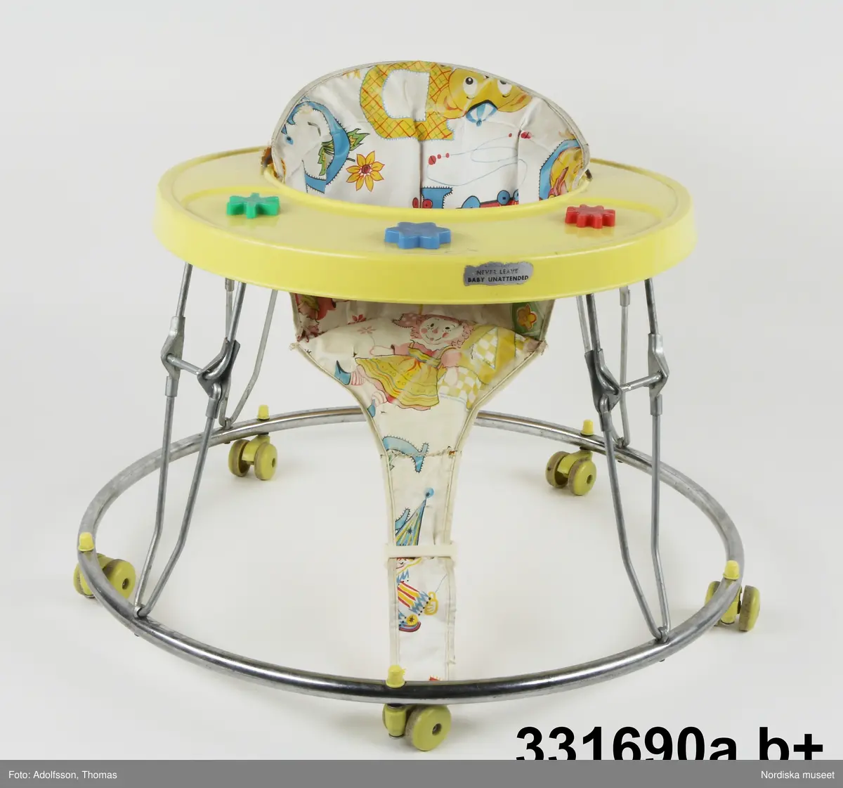 Gångstol (a) med lös sits (b).

a) Gångstol (lära-gå-stol) från 1960-talet, tillverkad i Taiwan. Ställning av metall, fem små hjul och aktivitetsbord av gul hårdplast med aktivitetsknappar i klara färger som barnet kan snurra eller trycka på. Den bärande metallställningen är hopfällbar.

b+) Den avtagbara sitsen av mönstrad pvc-plast är vadderad med skumgummi. Den har sprickor och är lagad med stygn på några ställen. Mönstret avbildar leksaker som teddybjörn, anka, docka och tåg samt bokstäver mot vit botten. Undersidan gul. Sitsen sitter fast vid ställningen genom en bågformad metallten som löper genom  sitsens ryggparti. 

Ihopmontering av sits och bord: Metalltenen skjuts in i överkanten på sitsens ryggparti. Sitsen sätts i hålet på aktivitetsbordet och sitsens metallten fästen i de små hålen på sidorna av hålet i aktivitetsbordet samt i ett fyrkantigt hål baktill på aktivitetsborden med hjälp av ett metallbleck som omsluter metalltenen.  Den smala framdelen/fliken av sitsen träs runt metallpinnen som sitter på aktivitetsbordets undersida och fästes genom plastspännet.

Anm. Nött och delvis något solkig. Sitsen är lagad med stygn på ett par ställen.
/Karin Dern 2012-11-15