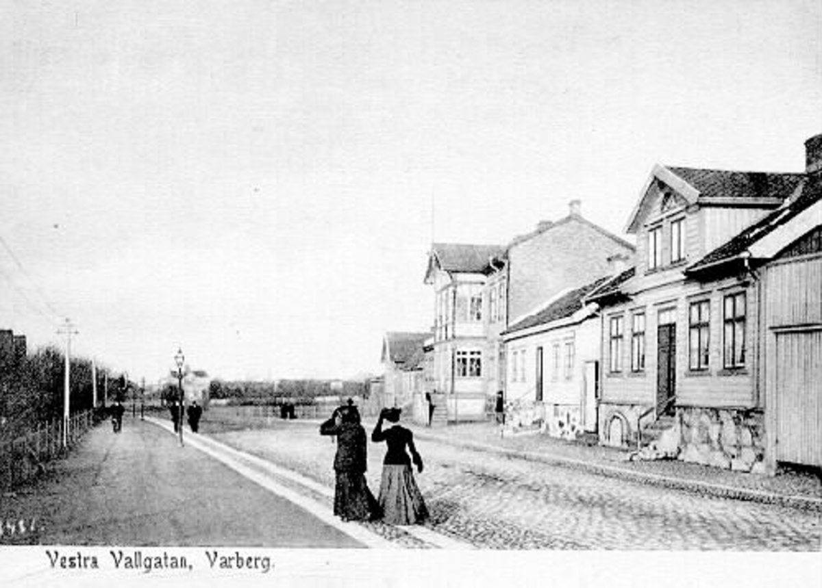 Brevkort, "Vestra Vallgatan, Varberg" (meddelandet skrivs på den tomma ytan på framsidan). Bilden är tagen mot norr så till höger syns kvarteret Berget och längs vänster sida ligger Järnvägsparken. Två damer promenerar och håller i hattarna. De vita stolparna till höger är Hugo Gerlachs elstolpar (uppfördes 1892) som går från hans Mekaniska verkstad, via Järnvägs- och Engelska parkerna till hans bostad i Franckalyckan. Stadens gatlyktor står på gångstråket.