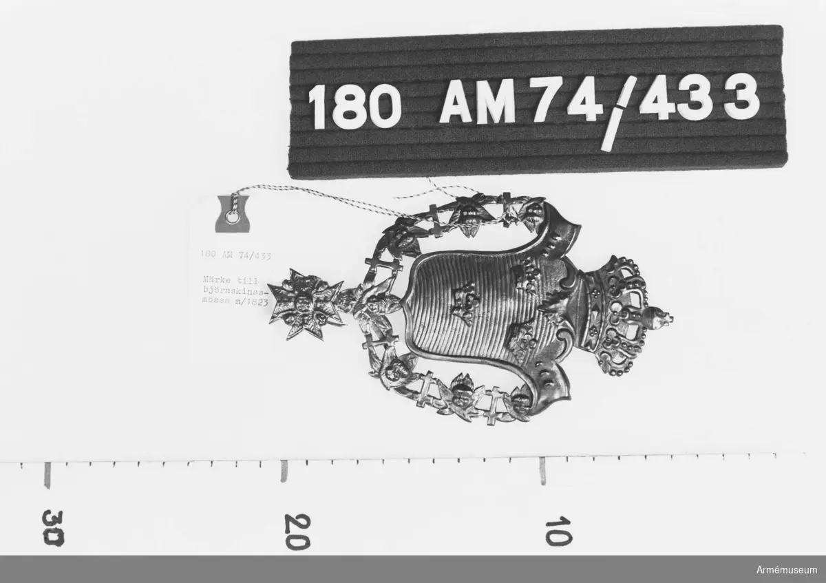 Förgylld plåt med lilla riksvapnet, serafimerkedja och kunglig krona.
