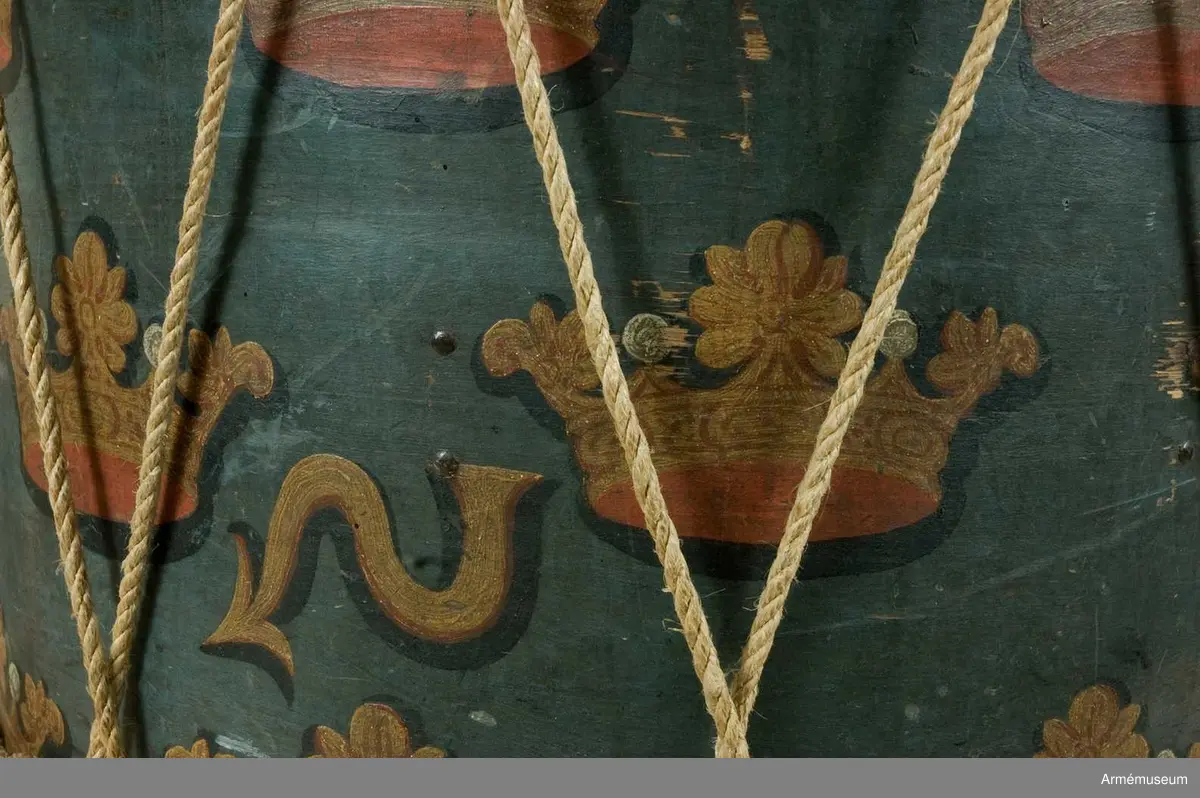 Regementstrumma från Fredrik I:s tid.
Blåmålad med sjutton st. strödda och öppna kronor, 90 mm höga. Fredrik I:s spegelmonogram krönt med sluten krona. Trumman är hopnitad till höger om monogrammet med 21 st nitar. 70 mm från skarven ett resonanshål. Hålet är omgivet av 17 nitar. Som dekor? Hopskarvningen går 190 mm omlott och även här nitat med nio st nitar. 
Snickarens bomärke "PP" finner man på den krona som befinner sig överst vid skarven. Mot höger finner man strax nedanför mitten en "S"-formad pil. Troligen ett skvadron- eller kompanimärke. (G K). Två nitar vid pilens bas som fasthåller en träförstärkning på insidan, inalles tolv st. Ytterligare en förstärkning högre upp något smalare, med den verkar endast fastlimmad. Nedtill finns en förstärkning och här finns hål och rester av gamla spikar. Stommen har varit försedd med band upp- och nedtill i sitt ursprungliga skick.