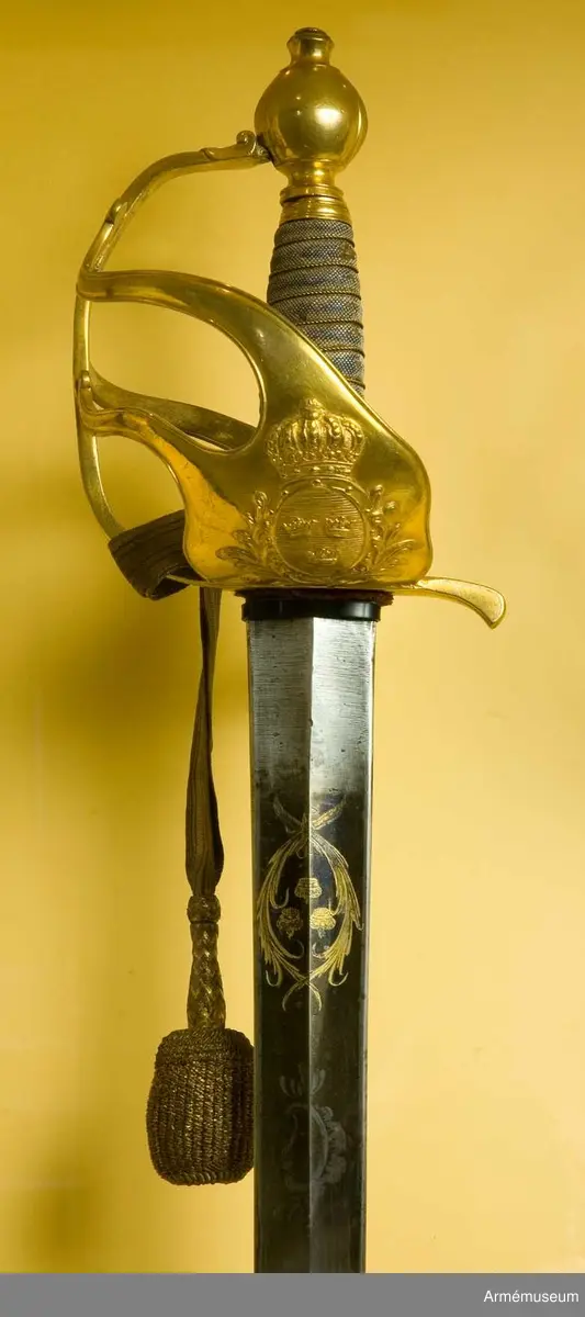Fäste av förgylld mässing med två yttre och en inre sidobygel. Parerplåten har en krönt sköld omgiven av stiliserade palmkvistar. Kaveln klädd med hajskinn och lindad med en tvinnad,  förgylld mässingtråd. 

Klingan är rak och tveeggad. Bredd vid fästet: 37 mm. Upptill rester av blånering och dekorerad med tre kronor, omslutna av två palmkvistar.
