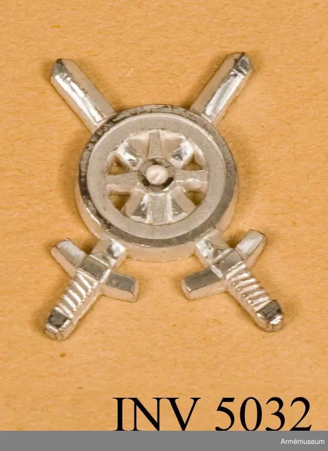 Tillverkat av metall. I likhet med emblemen till  trängtruppernas äldre uniformer är det här utfört i silverfärg. Samma motiv som i 1939 års modell, ett kärrhjul på två korslagda svärd, men något annorlunda utformat.
